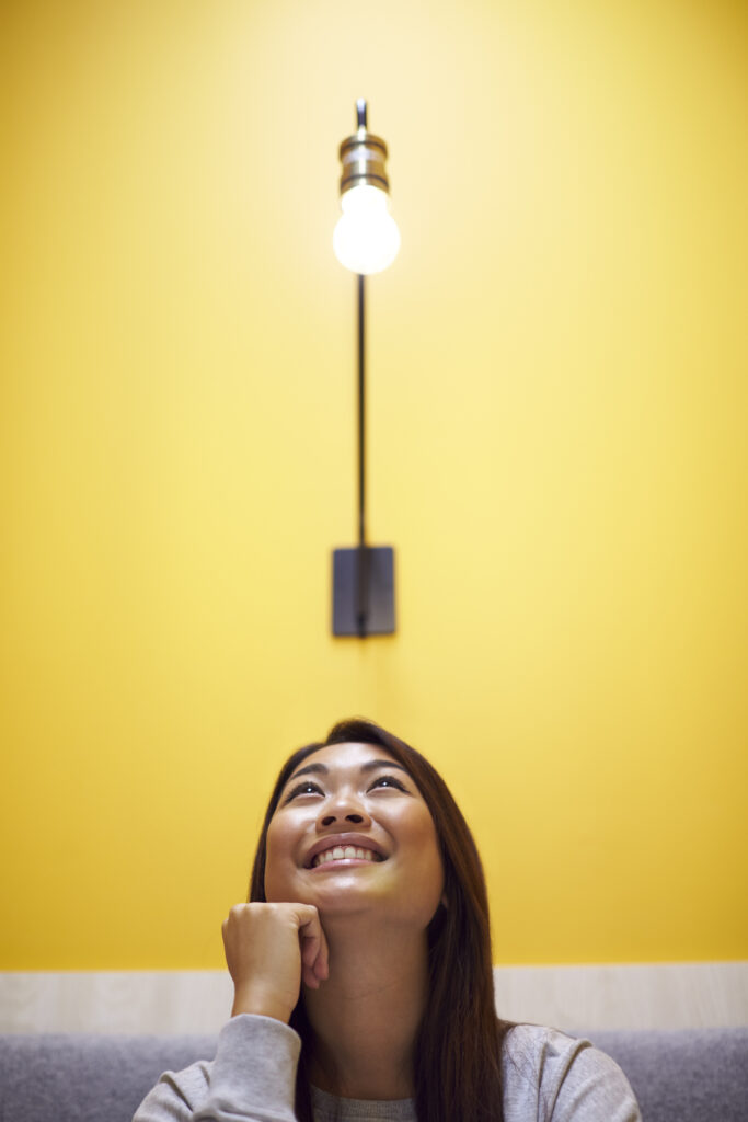 Woman Sitting Under Light Bulb In Office Suggesting Inspiration Or Idea